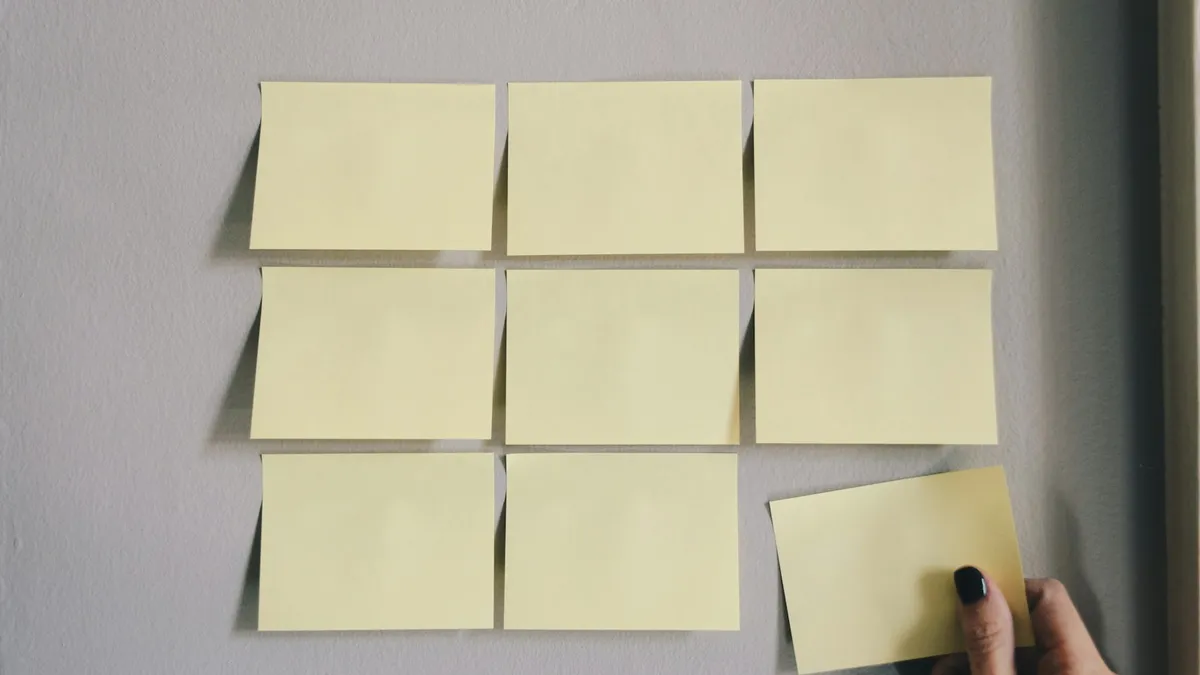 Main posant un post-it jaune sur un mur, à côté d'une grille de huit post-it vierges alignés